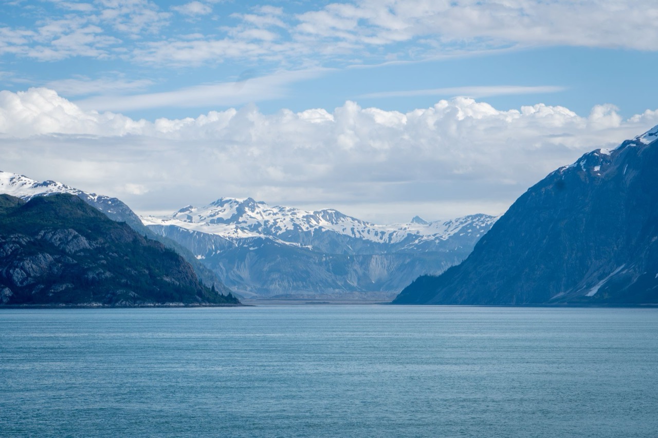 Snow-streaked mountains opening onto still water in an Alaskan fjord, under a wide, clouded sky